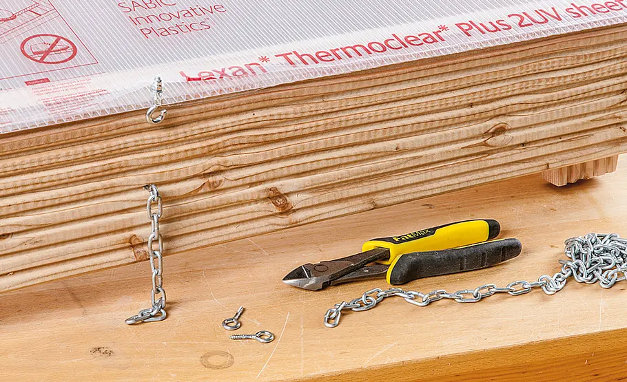 Eine Kunststoffplatte ist mit Ketten und Schraubhaken am Holz befestigt, ein Seitenschneider und Ketten liegen vorne.