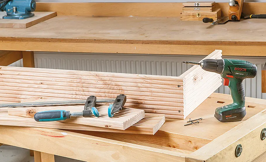 Holzbretter werden mit Schrauben zu einem Kasten zusammengeschraubt, Schraubzwingen und Werkzeug liegen auf der Werkbank.