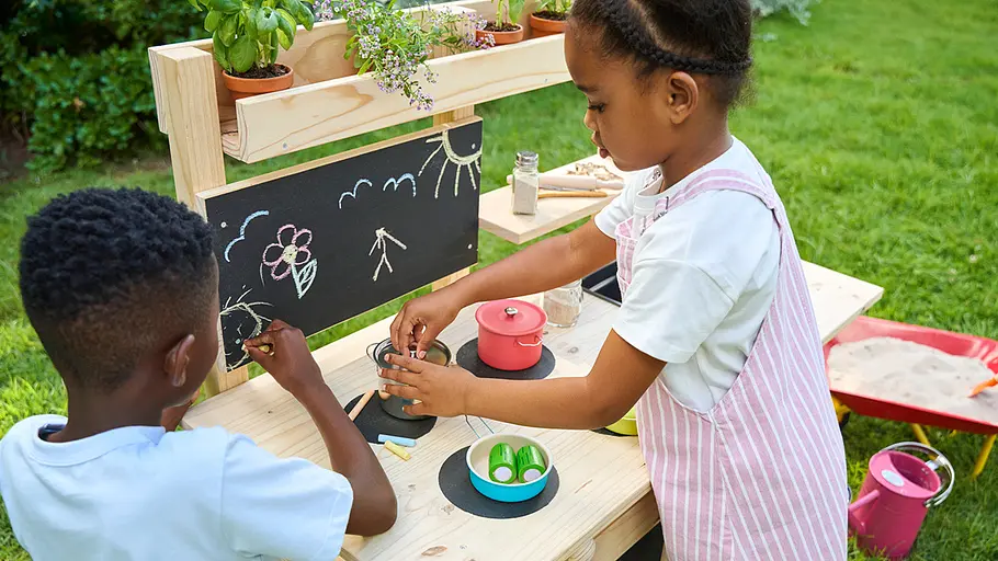 Matschküche: Fertig! Zwei Kinder spielen an der fertigen Matschküche im Garten, malen mit Kreide und kochen mit Töpfen auf der Holzarbeitsfläche.