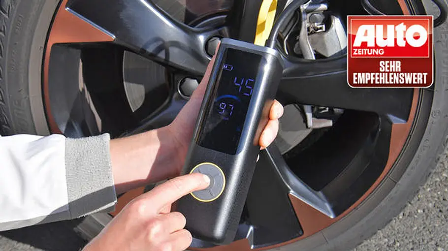 Eine Person hält eine schwarze Fanttik-Akku-Luftpumpe mit leuchtendem Display vor einem Autoreifen; Empfehlungssiegel sichtbar.