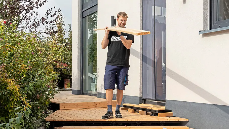 Terrasse gestalten Ein Mann trägt ein Holzbrett auf der Schulter und steht auf einer teilweise gebauten Holzterrasse neben einem modernen Haus.