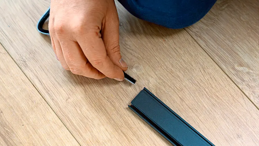 Eine Hand steckt eine schwarze Bürstendichtung in einen Metallrahmen auf einem hellen Holzfußboden für eine Insektenschutztür.
