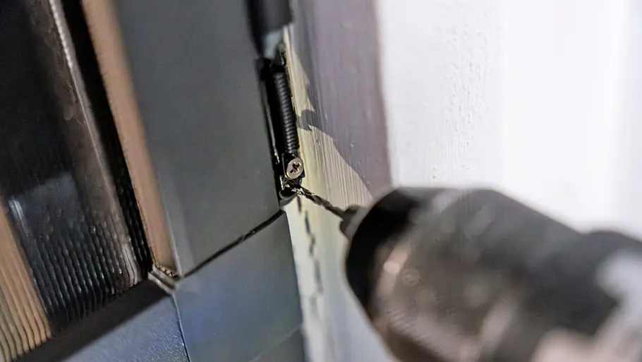 Eine Bohrmaschine zieht eine Schraube am Scharnier mit Feder einer Insektenschutztür an einen weißen Türrahmen fest.