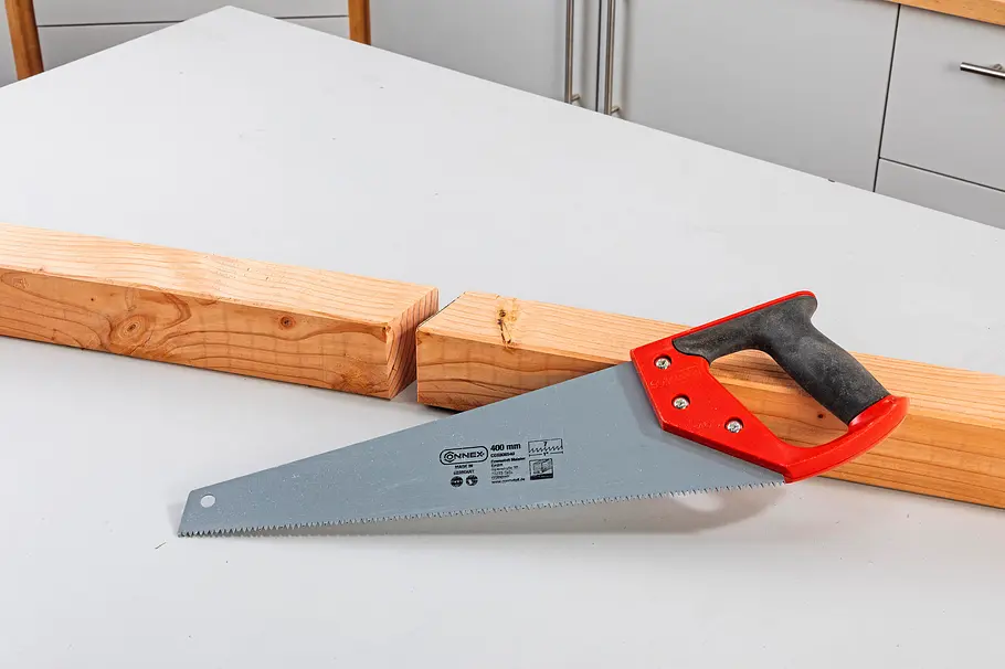 Gartendusche bauen Eine Handsäge mit rotem Griff liegt vor einem langen Holzbalken mit eingesägter Aussparung.