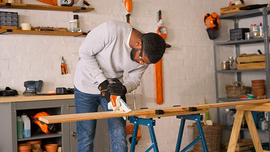 Ein Mann in einer Werkstatt schneidet mit einer Säge ein Holzbrett, das auf zwei Böcken gespannt ist.