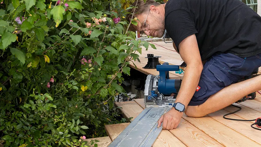 Terrasse gestalten Ein Mann schneidet die überstehenden Terrassendielen am Terrassenrand mit einer Handkreissäge und Führungsschiene ab.