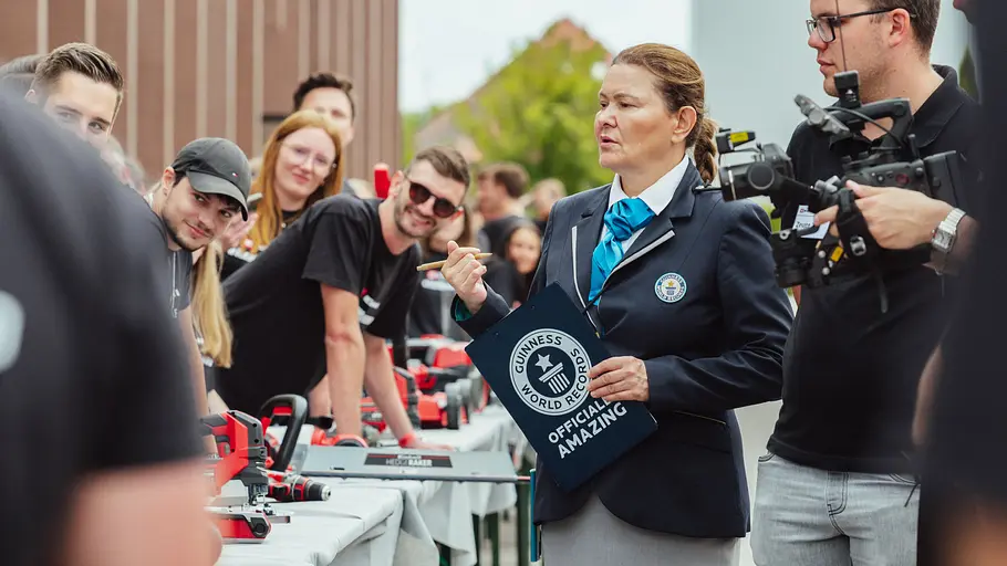 Guinness World Records-Jurorin mit Klemmbrett und offiziellem Abzeichen steht zwischen Teilnehmenden und beobachtet die Aktion am langen Tisch mit Einhell-Geräten.