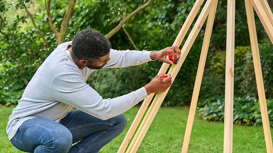 Ein Mann misst mit Maßband und Bleistift die Höhe einer Holzlatte am aufgebauten Tipi-Gestell im Garten ab.