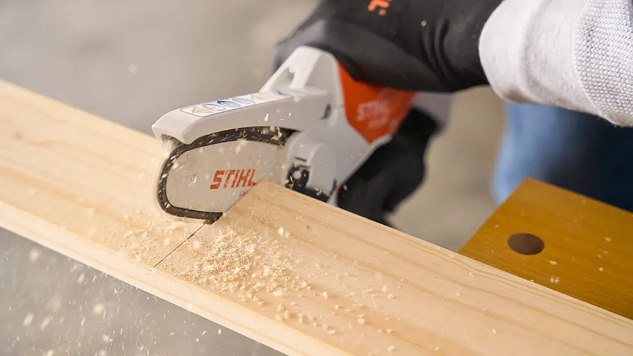 Matschküche: Latten zusägen Eine Person schneidet mit einer STIHL-Kettensäge eine Holzlatte auf einer Werkbank entlang einer Markierung ab.