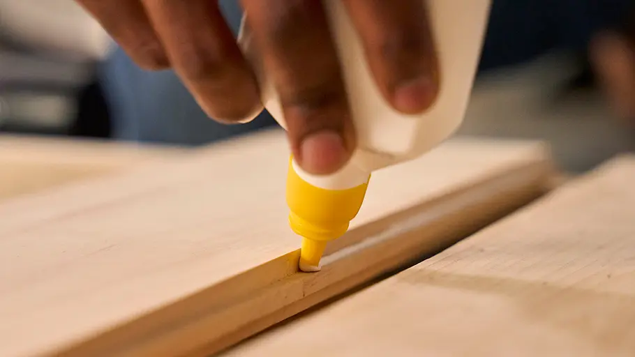 Eine Hand trägt mit einer Flasche Holzleim auf die Kante eines Holzbretts auf, weitere Bretter liegen daneben.