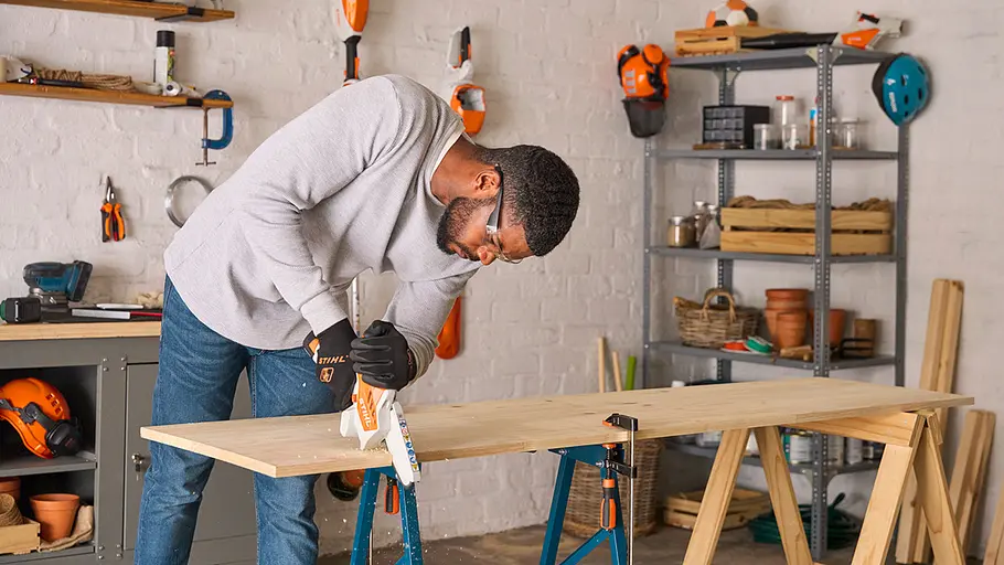 Matschküche: Arbeitsplatte Ein Mann sägt eine große Holzplatte, die auf zwei Sägeböcken liegt, als Arbeitsplatte für die Matschküche zurecht.