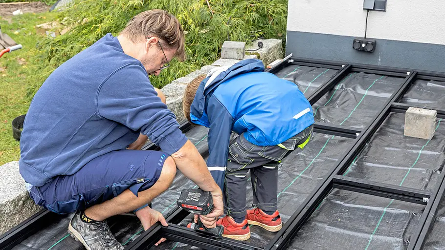Terrasse gestalten Ein Mann und ein Kind verschrauben gemeinsam das Metalluntergestell auf der vorbereiteten Terrasse mit Unkrautvlies im Garten.