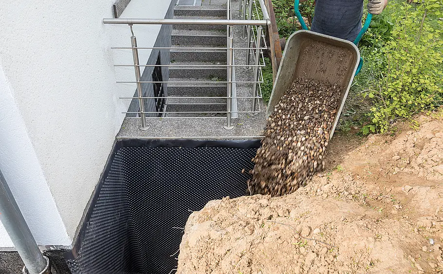 Auffüllen der Baugrube mit Kies an abgedichteter Hauswand Eine Person schüttet mit einer Schubkarre Kies auf eine mit Noppenfolie geschützte Baugrube an der Hauswand.