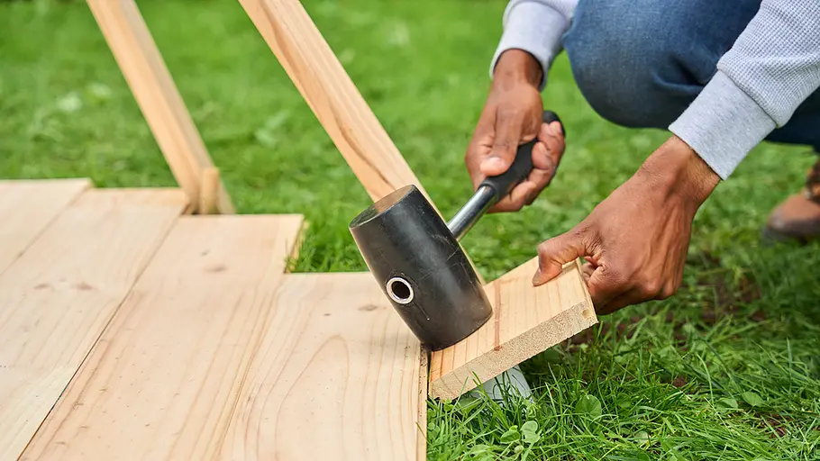 Eine Person hämmert mit einem Gummihammer ein Holzstück an der Kante einer Holzbodenplatte ins Gras.