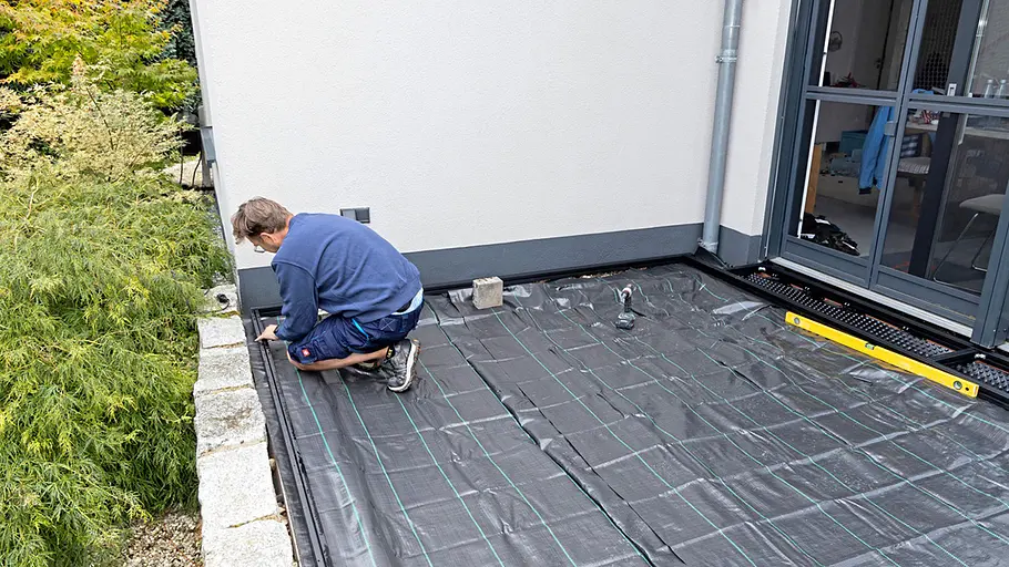 Terrasse gestalten Ein Mann kniet auf einer mit schwarzem Unkrautvlies bedeckten Terrasse vor dem Haus und arbeitet an der Vorbereitung des Untergrunds.