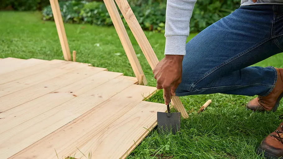 Eine Person kniet auf dem Rasen und gräbt ein Loch neben die Holzbodenplatte des Zeltes.