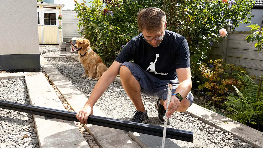 Terrasse gestalten Ein Mann misst auf einer Baustelle im Garten mit Zollstock und Wasserwaage, ein Hund sitzt im Hintergrund auf dem Kies.