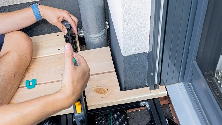 Terrasse gestalten Ein Mann richtet eine zugeschnittene Holzdiele mit einer Zwinge an einer Ecke mit Rohr und Terrassentür aus.