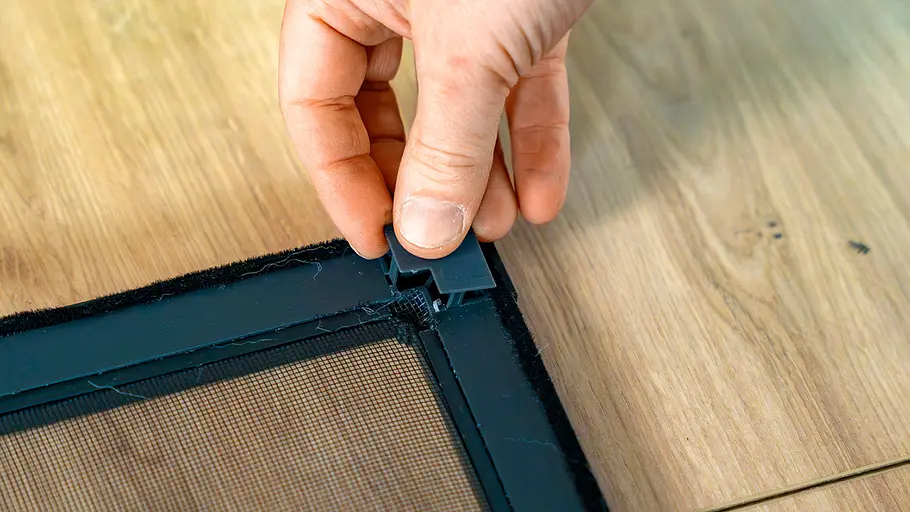 Eine Hand drückt ein kleines schwarzes Kunststoffteil auf die Ecke eines Insektenschutzrahmens mit Bürstendichtung auf Holzboden.