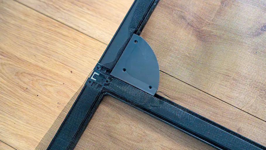 Der schwarze Metallrahmen mit feinem Netzgewebe liegt auf einem hellen Holzboden, Ecke mit Befestigungselement sichtbar.