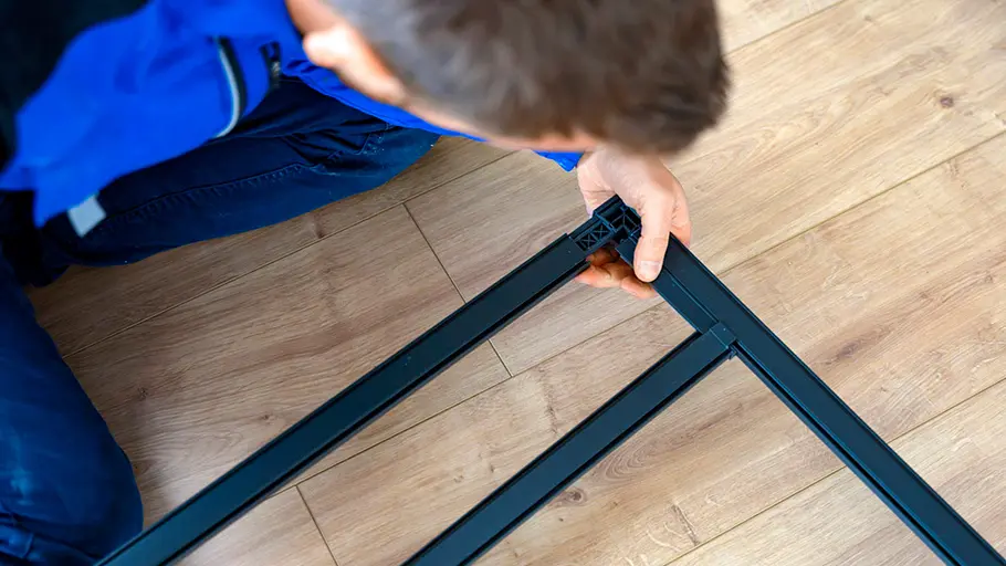 Eine Person in blauer Arbeitskleidung montiert auf einem Holzfußboden einen schwarzen Metallrahmen für eine Insektenschutztür.