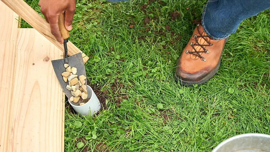 Mit einer Kelle werden kleine Steine in ein weißes Kunststoffrohr neben einer Holzbodenplatte auf einer Wiese gefüllt.