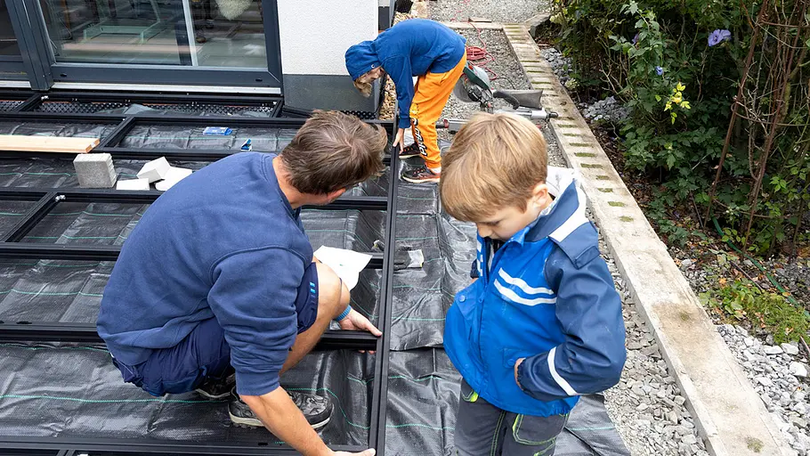 Terrasse gestalten Ein Mann richtet zusammen mit zwei Kindern die Metallunterkonstruktion für die Terrasse auf schwarzem Unkrautvlies aus.
