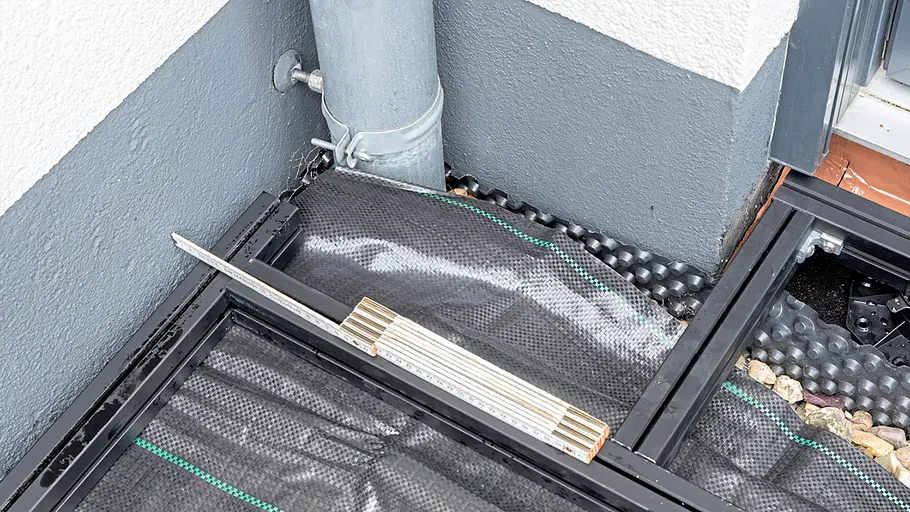 Terrasse gestalten Der Boden vor der Hauswand zeigt eine Unterkonstruktion mit schwarzem Unkrautvlies, Zollstock und Abflussrohr in einer Ecke.