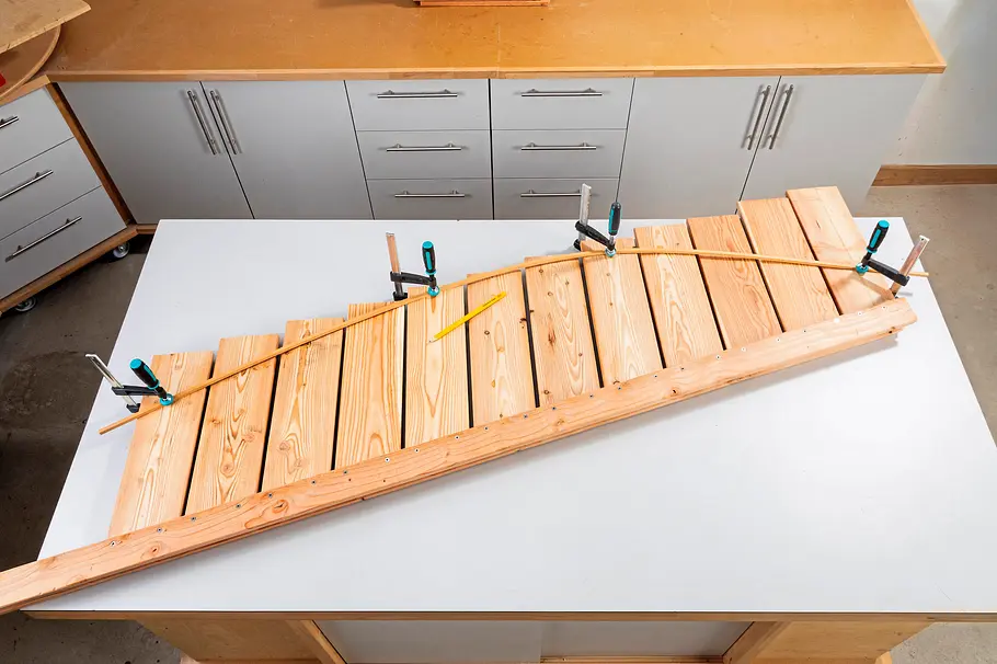 Gartendusche bauen Eine blattförmige Holzkonstruktion ist mit mehreren Schraubzwingen auf der Werkbank fixiert, während ein Kupferrohr darauf ausgerichtet wird.