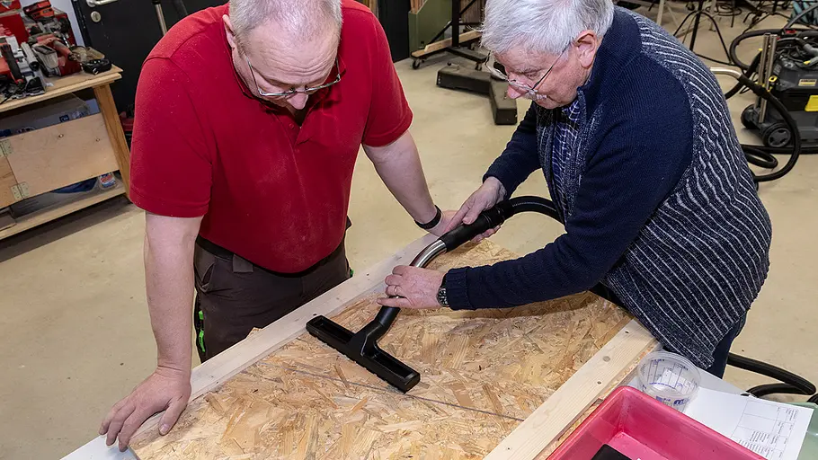 Werksattsauger Sand absaugen Zwei Männer reinigen gemeinsam eine Arbeitsplatte aus Holz mit einem Werkstattsauger in einer Werkstattumgebung.