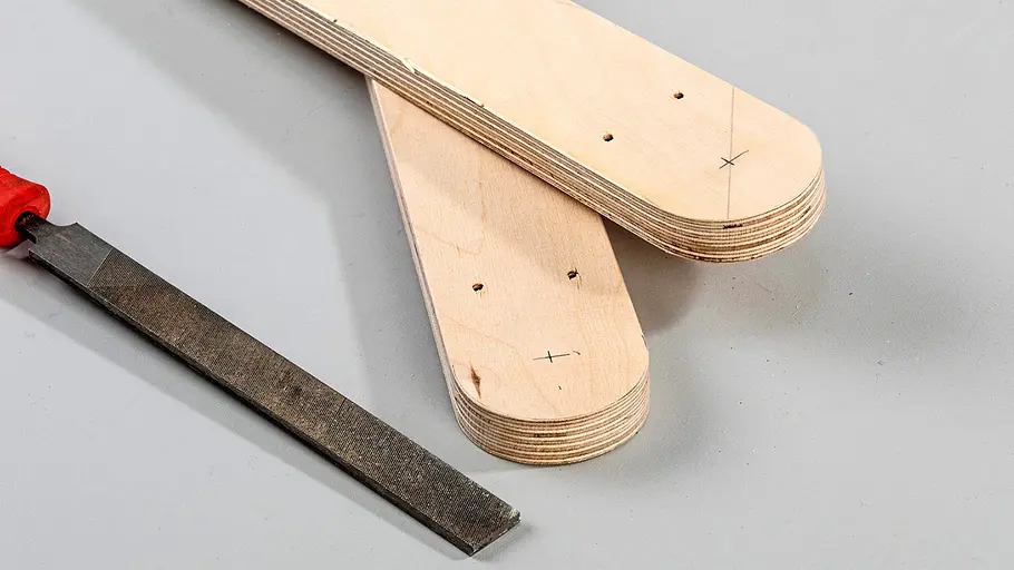 Abrunden mit Feile Zwei Holzleisten mit abgerundeten Enden und Bohrlöchern liegen neben einer Flachfeile mit rotem Griff auf grauer Fläche.