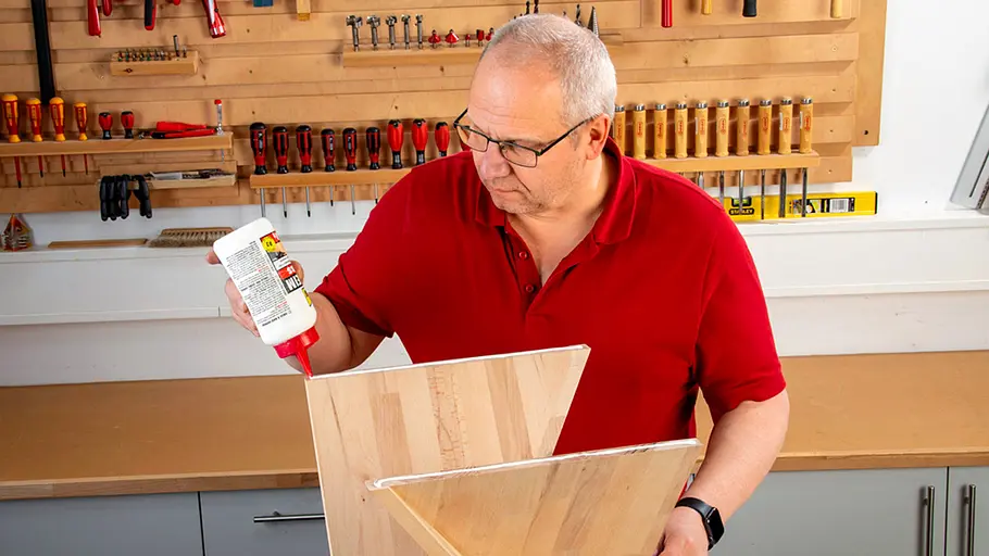 Verdrehter Hocker Ein Mann trägt in einer Werkstatt Holzleim auf die Kanten zweier schräg montierter Holzplatten auf.
