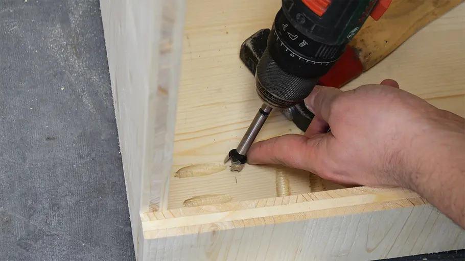Paketbox Eine Hand hält ein Holzbrett, während mit einem Akkuschrauber Verbindungslöcher in den Boden einer Paketbox gebohrt werden.
