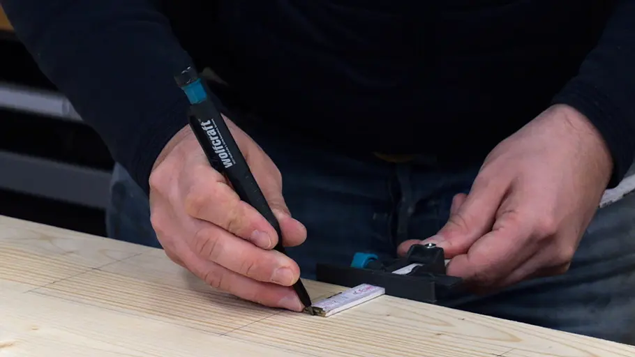 Paketbox Eine Person misst mit einem Zollstock und markiert mit einem Bleistift Maße auf einer hellen Holzplatte.