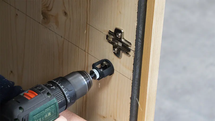 Paketbox Ein Akkuschrauber bohrt ein Loch in die Seitenwand einer Holz-Paketbox, neben einem bereits montierten Metallscharnier.