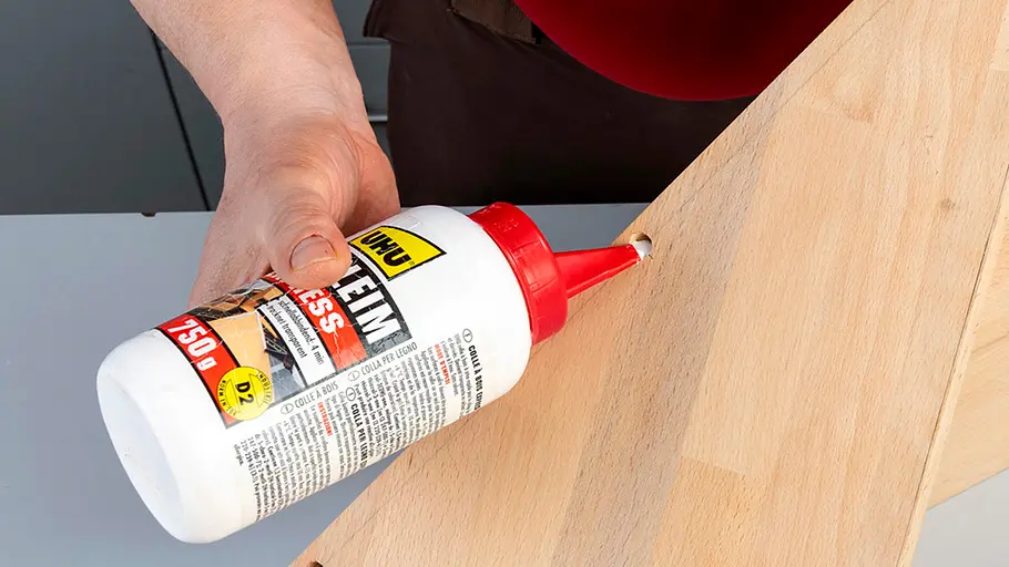 Verdrehter Hocker Eine Person füllt mit einer weißen Leimflasche Holzleim in ein gebohrtes Loch einer Holzplatte.