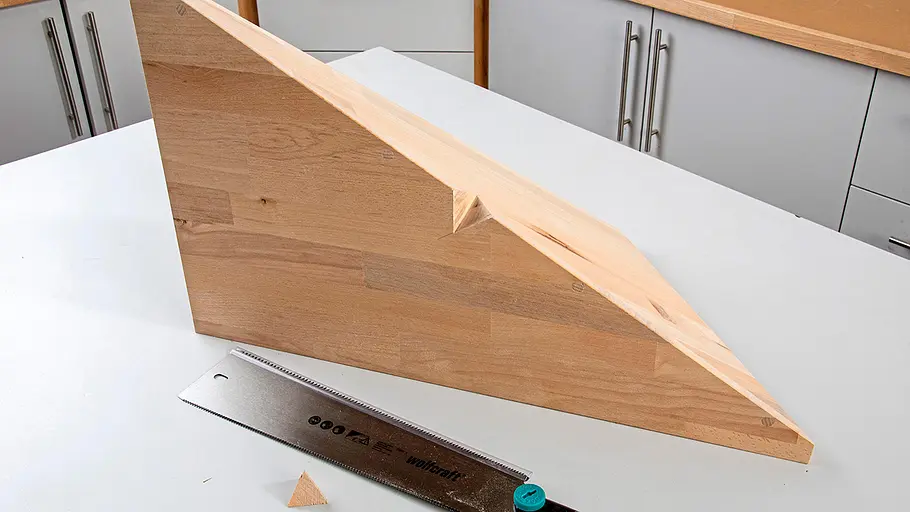 Verdrehter Hocker Ein Holzbrett mit schräger Schnittkante liegt auf einem Tisch, davor eine Handsäge und ein ausgeschnittenes Holzdreieck.