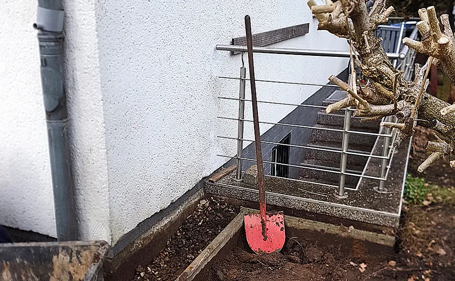 Freigelegte Hauswand mit Spaten vor Abdichtung Ein Spaten mit rotem Blatt steht an einer freigelegten Hauswand neben einem Busch und einem Metallgeländer.