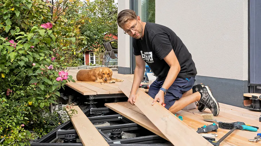 Terrasse gestalten Ein Mann verlegt Terrassendielen, während ein Hund entspannt auf den bereits verlegten Brettern in der Sonne liegt.