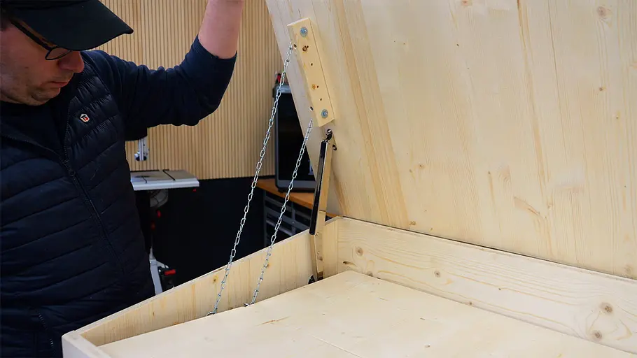 Paketbox Eine Person hält den Deckel einer großen Holzkiste, der mit einer Kette und einem Metallbeschlag innen gesichert ist.