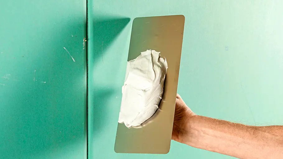 Spachtelmasse Weiße Spachtelmasse ist auf einer rechteckigen Glättkelle verteilt, die vor einer grünen Wand gehalten wird.