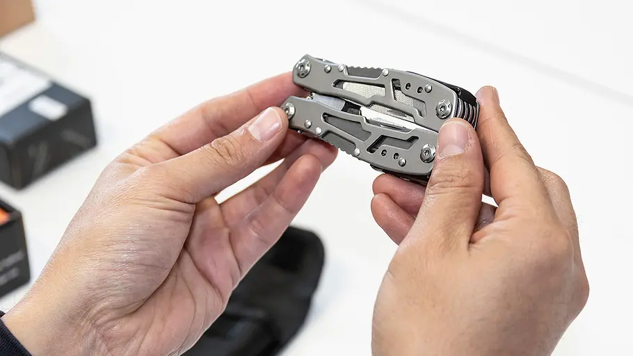 Multitool-Test Zwei Hände halten ein silbernes Multitool in geschlossener Form über einem weißen Tisch mit Verpackungen.