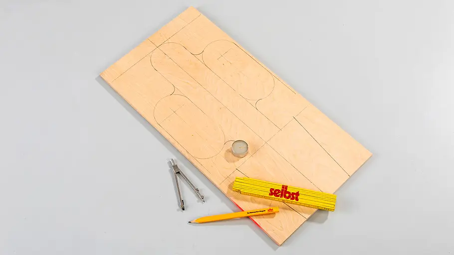 Katzen-Kratzbrett: Vorarbeit Ein Holzbrett mit aufgezeichneten Umrissen, daneben ein Bleistift, ein Zirkel und ein Zollstock auf grauer Fläche.