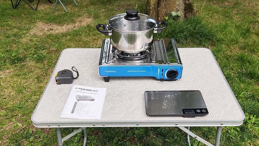 Campingaz Camp`Bistro Ein blauer Campingkocher mit Edelstahltopf steht mittig auf einem Campingtisch, daneben liegen eine Waage, ein Maßband und eine Anleitung.