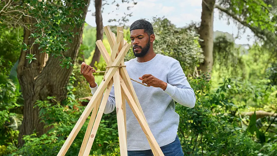 Ein Mann steht auf einer Wiese und verknotet eine Kordel um die Spitzen von aufgestellten Holzlatten zu einem Tipi-Gerüst.