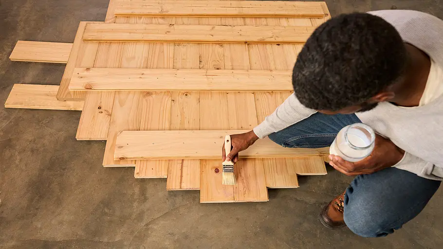 Ein Mann sitzt auf dem Boden und streicht eine zusammengesetzte Holzfläche mit einem Pinsel und einer Dose Holzschutzmittel.