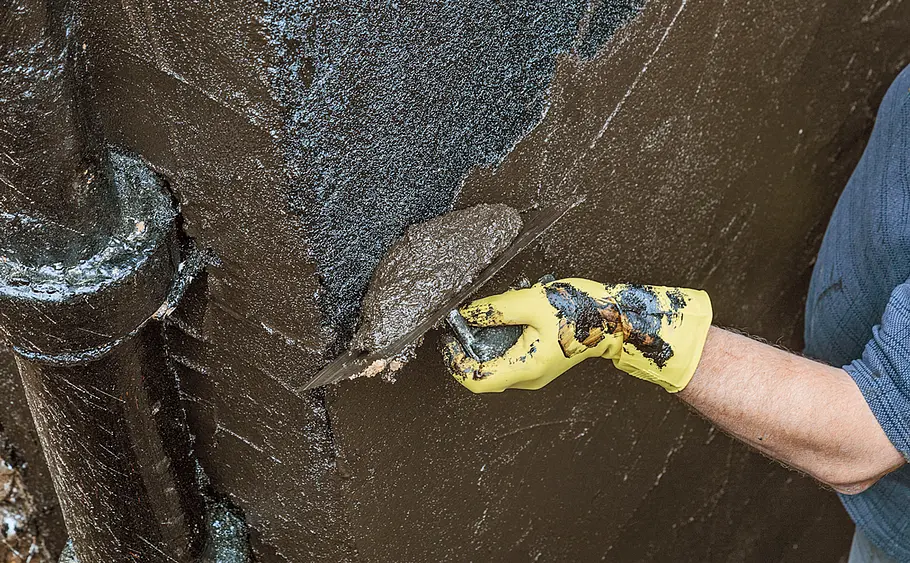 Hauswand mit Bitumen abdichten Mann trägt Bitumendickanstrich mit einer Kelle auf die freigelegte Kellerwand auf.