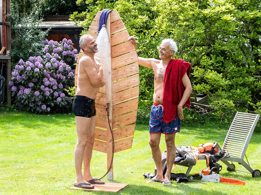 Gartendusche bauen Zwei Männer in Badebekleidung testen eine blattförmige Gartendusche aus Holz.