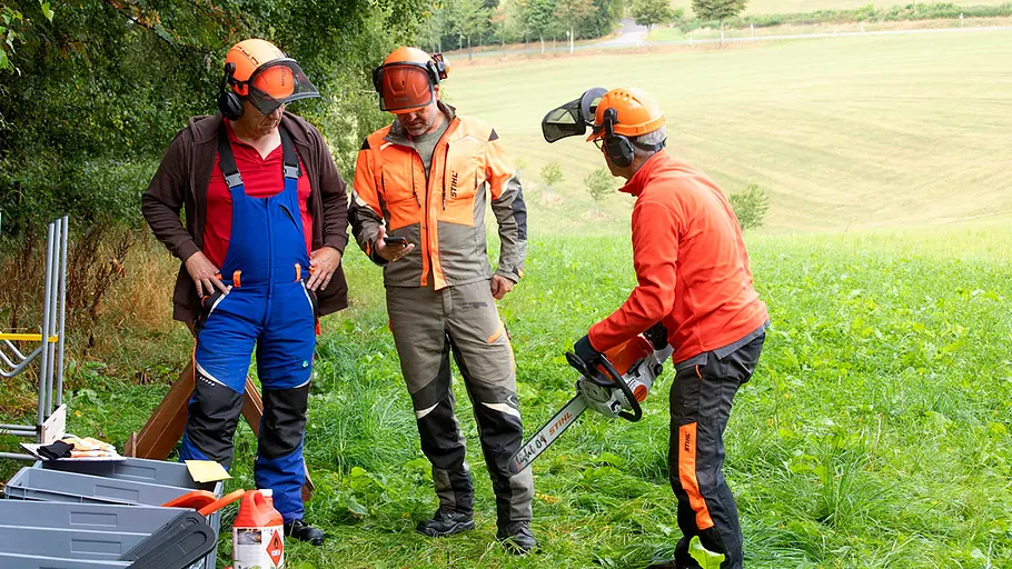 Erfahrunngsaustausch beim Motorsägen-Test Drei Personen mit Schutzkleidung stehen auf einer Wiese, eine hält eine Motorsäge, die anderen schauen auf ein Smartphone.
