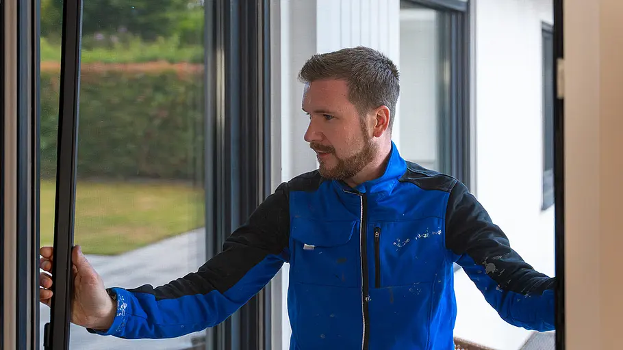 Eine Person in blauer Arbeitskleidung baut eine Insektenschutztür an einer großen Terrassentür in einem modernen Haus ein.